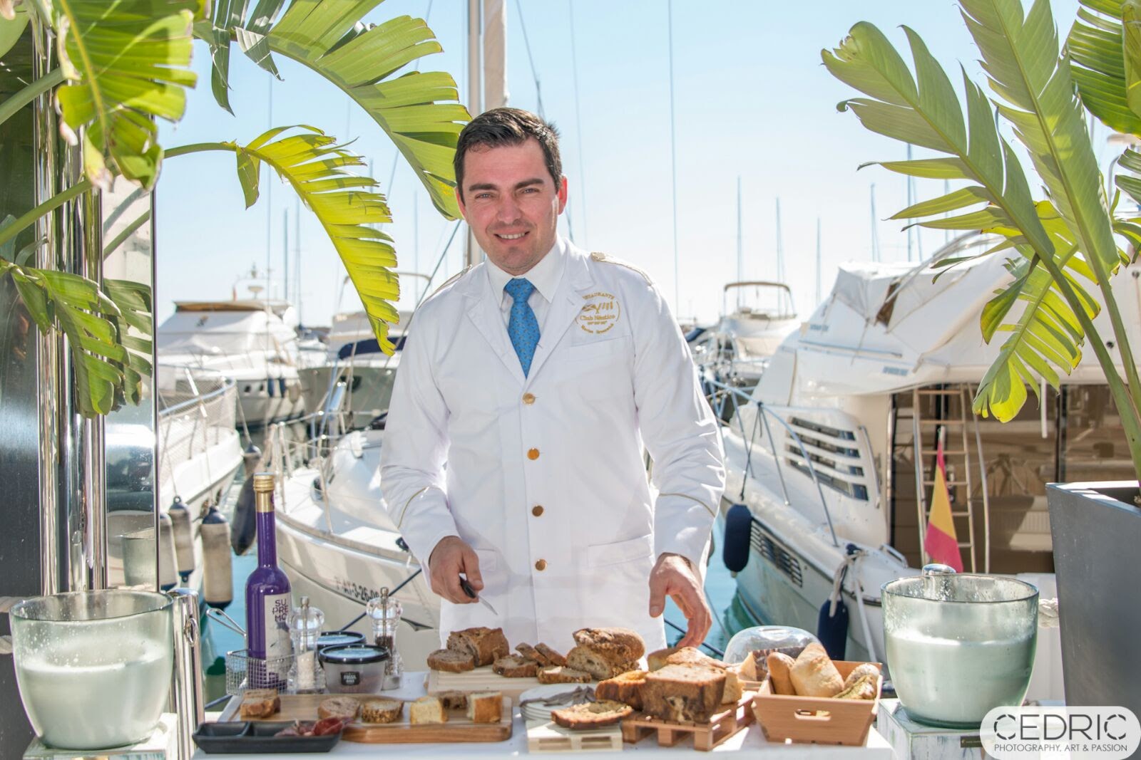 Mario Andries con chaquetilla blanca y corbata azul cortando pan artesano en una mesa con panes y aceite de oliva, frente a los veleros de un club náutico. Fotografía de Cedric.